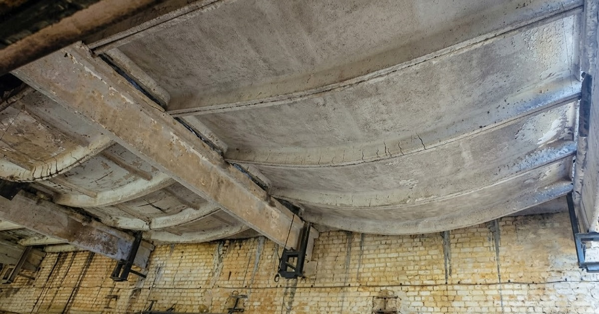 Interior view of a reinforced concrete solid slab with beams showing visible structural deflection and sagging in an industrial setting.